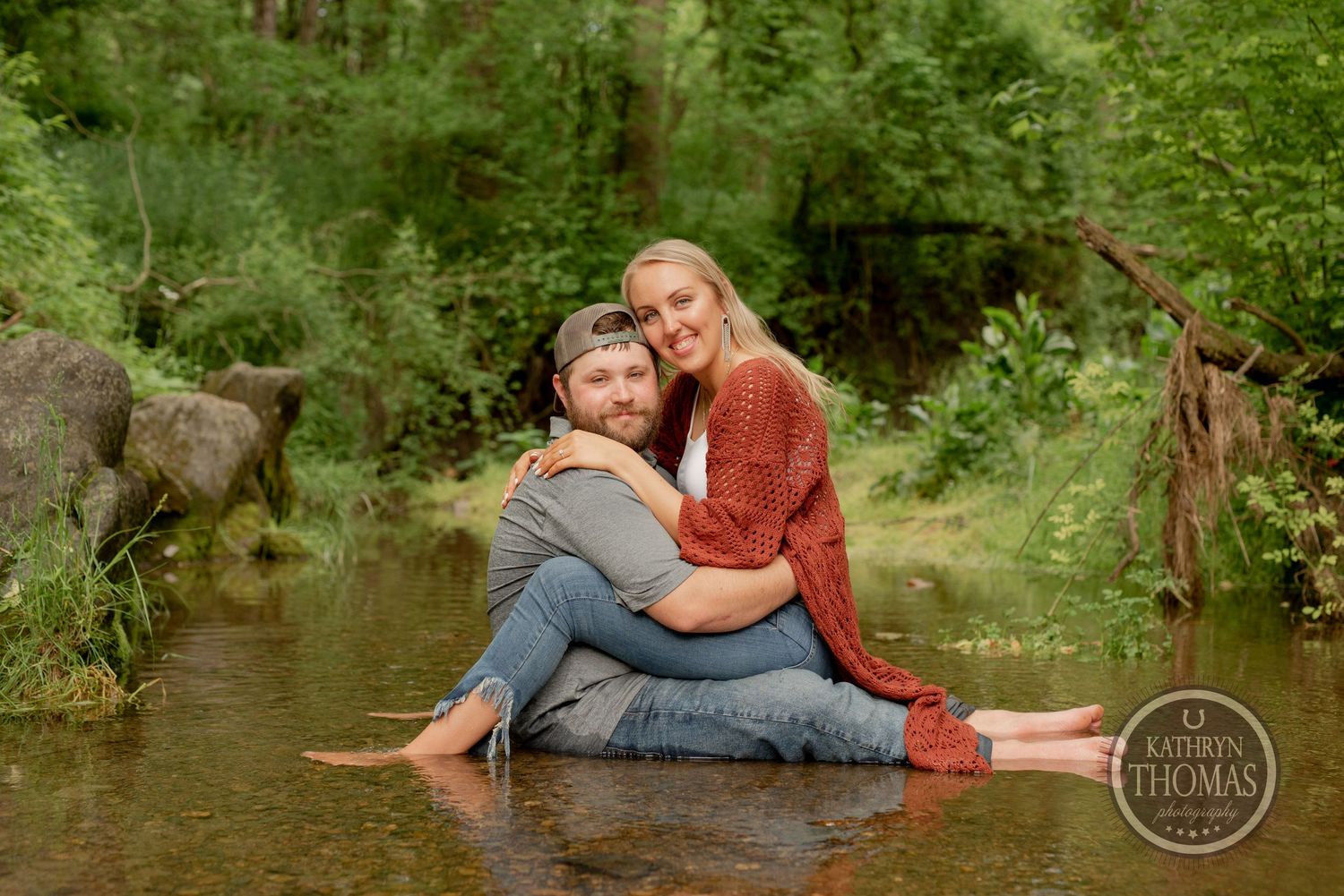 A couple sits together intimately on rocks in a creek surrounded by green forest.