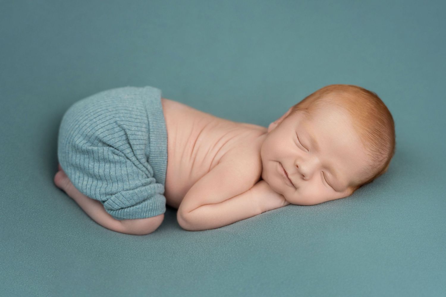 Sleeping newborn baby wearing blue shorts on teal background.