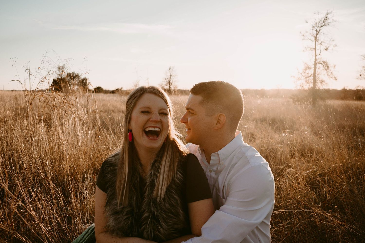A joyful couple shares a genuine laugh while sitting together in a sunlit field.