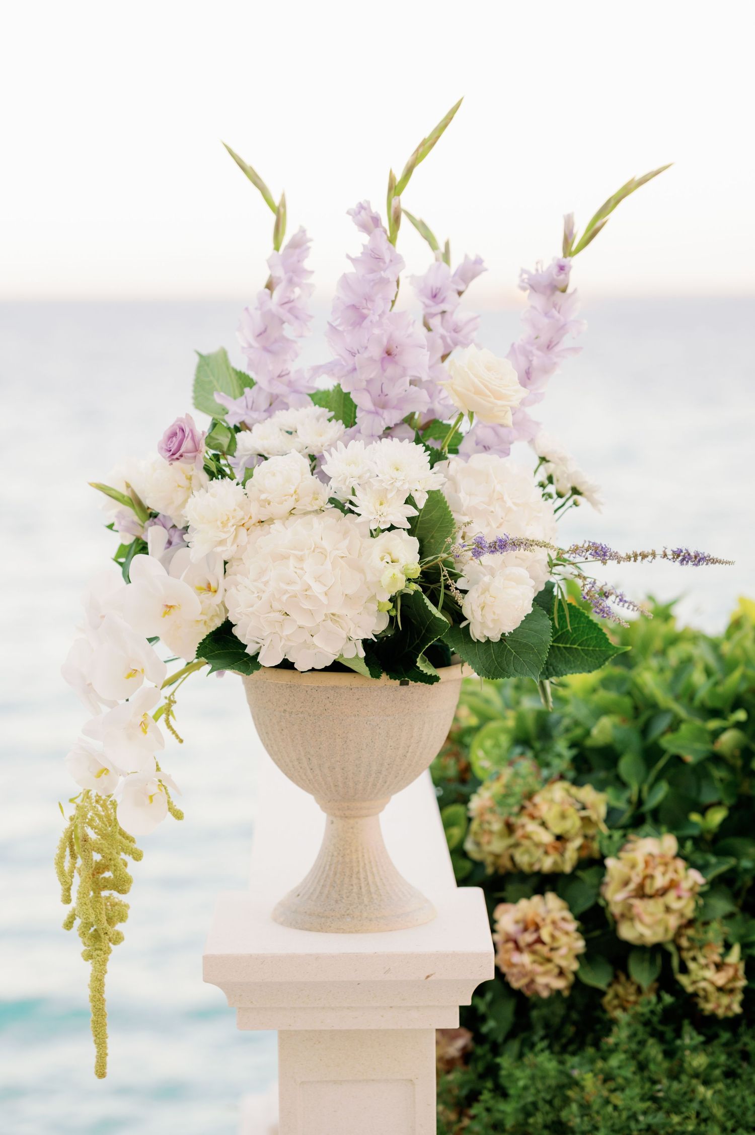 Elegant floral arrangement with white and lavender blooms in a classic stone urn vase against an ocean backdrop.