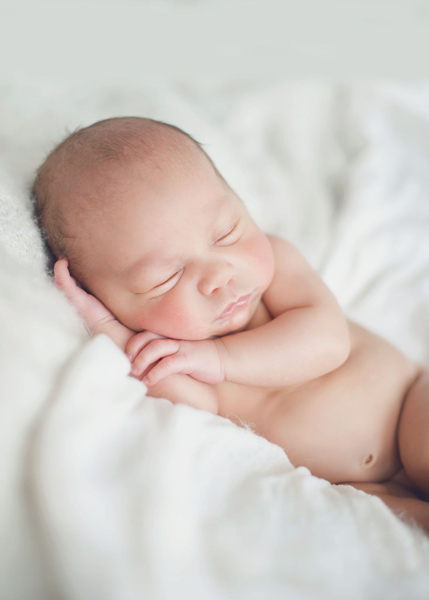A peaceful sleeping newborn baby rests on soft white bedding in natural light.