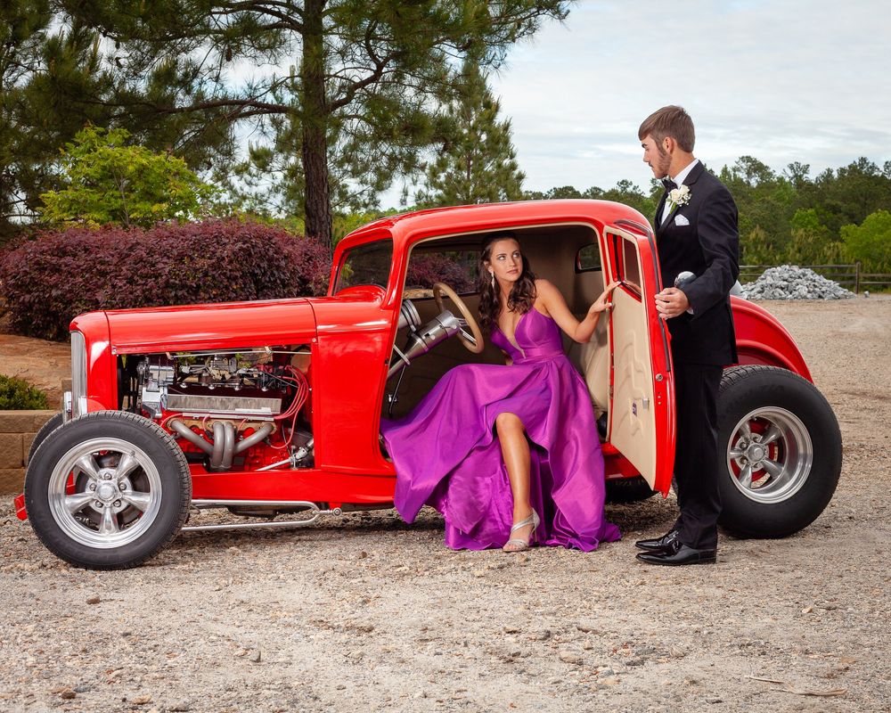 A bright red hot rod classic car with a purple-dressed passenger being photographed for a formal occasion.