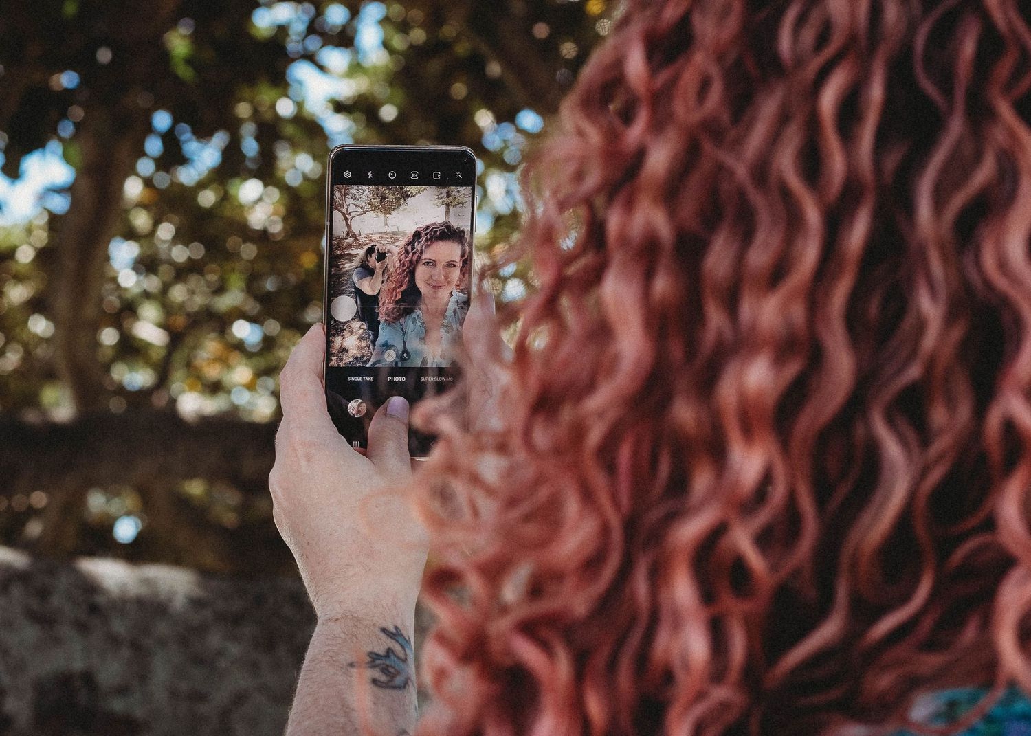 Hand holding phone taking selfie photo with curly hair visible in foreground.