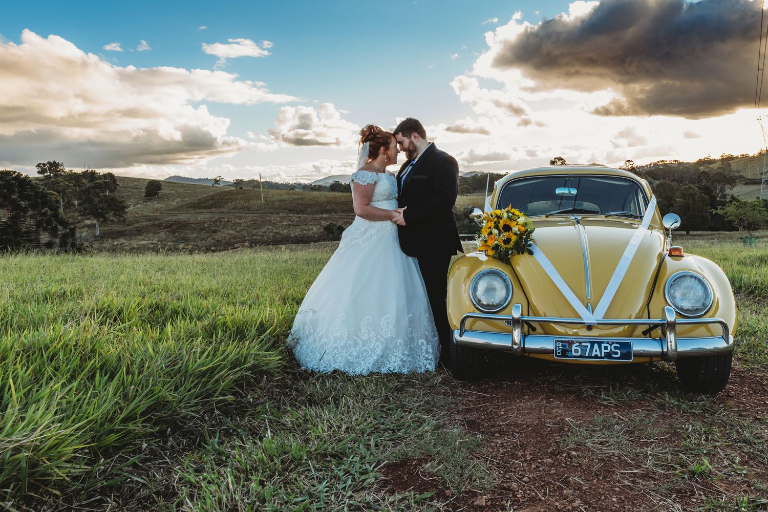 Wedding couple poses beside a classic yellow Volkswagen Beetle in a scenic field.