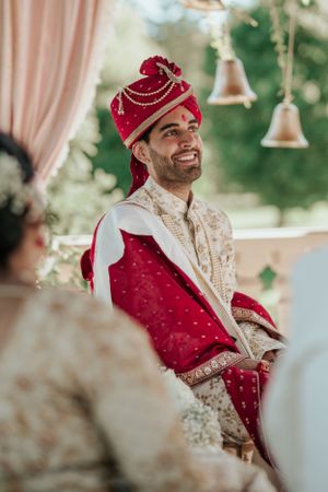Traditional Indian groom wearing red and cream wedding attire with ornate turban during ceremony.