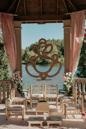 Intimate wedding ceremony setup inside a gazebo with pink drapes and decorative gold emblem overlooking water.