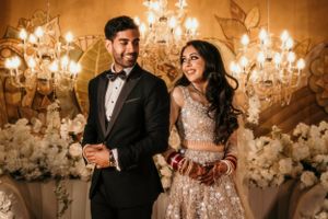 A glamorous couple poses against an elegant backdrop with chandeliers and white floral decorations at a wedding reception.
