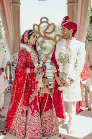 Indian wedding couple in traditional red and cream attire under decorated mandap.