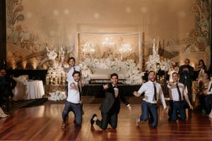 Groomsmen performing choreographed dance routine at wedding reception with decorated backdrop.