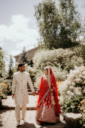Wedding couple in traditional South Asian attire posing in sunlit garden with blooming flowers.
