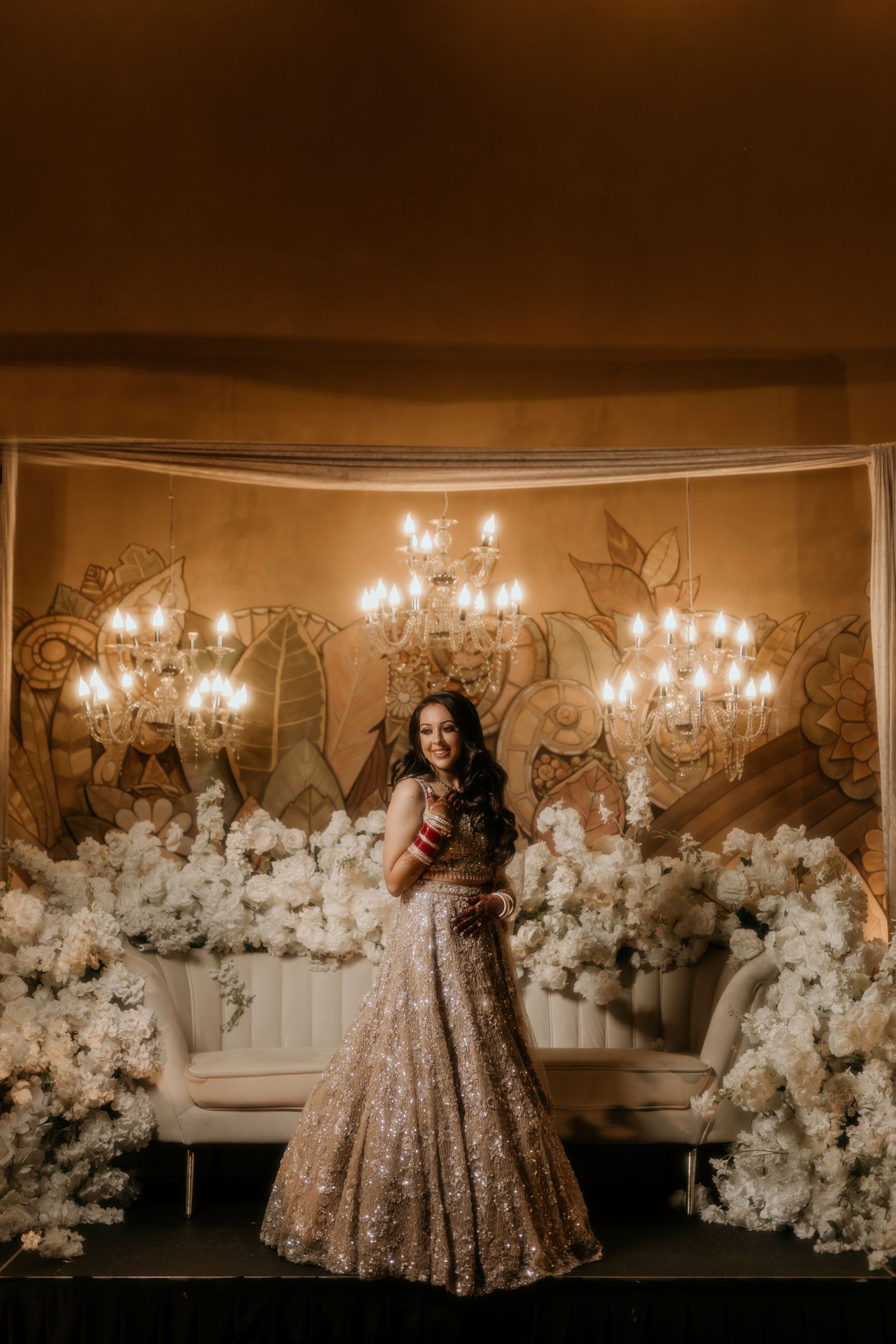A sparkling golden lehenga catches the light beneath elegant chandeliers and floral decor.