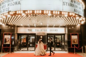 Couple in formal attire poses outside the Eglinton Grand theater with vintage marquee lighting.