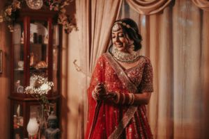 A bride in a red and gold embroidered lehenga stands elegantly against draped curtains.