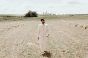 A figure in traditional white attire stands alone in an expansive rural field under overcast skies.