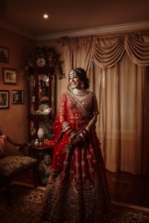 A traditional Indian bride in red poses gracefully in a warmly lit room with elegant decor.