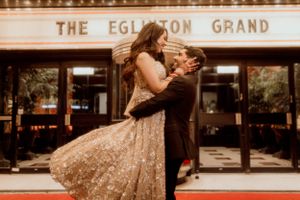 A romantic couple embracing in front of The Eglinton Grand theater entrance.