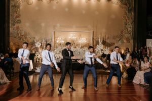 Groomsmen perform synchronized dance moves at an elegant wedding reception with ornate backdrop decor.