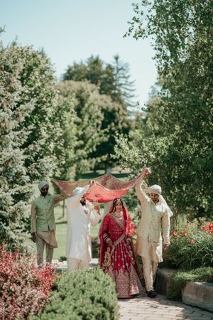 Garden wedding ceremony with red dupatta canopy held over couple walking through flowering path.