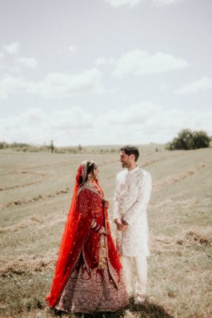 Indian wedding couple in traditional dress standing in open field under cloudy sky.
