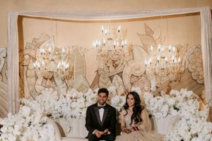 Wedding couple seated on ornate loveseat with white floral decorations and crystal chandeliers in background.