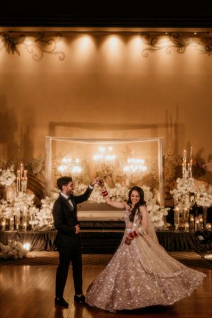 Couple dances in an elegantly lit ballroom decorated with white flowers and chandeliers.