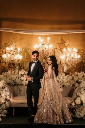 Couple in formal attire stands in front of illuminated chandeliers and white floral arrangements at an elegant event.