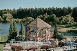 Beautiful lakeside gazebo decorated with pink floral arrangements and white chairs set up for an outdoor wedding ceremony.
