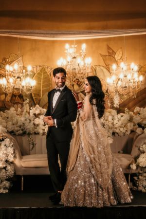 Elegant couple in formal wear poses against a backdrop of crystal chandeliers and white flowers.