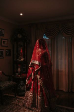 A traditional red and gold bridal lehenga displayed dramatically in a dimly lit room with draping curtains and framed artwork.