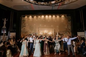 Wedding guests and wedding party dancing together in a large reception hall with decorative lighting.