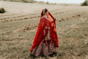 An ornate red bridal lehenga with intricate embroidery billows in the wind on rolling hills.
