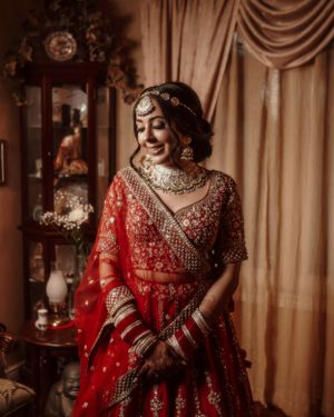 A bride wearing an intricately detailed red lehenga poses beside an ornate display cabinet.