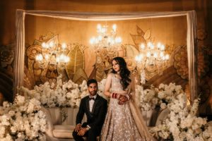 Wedding couple poses in front of a romantic backdrop with chandeliers and white floral arrangements.