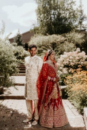 Traditional South Asian wedding couple in ornate white and red attire standing in garden setting.