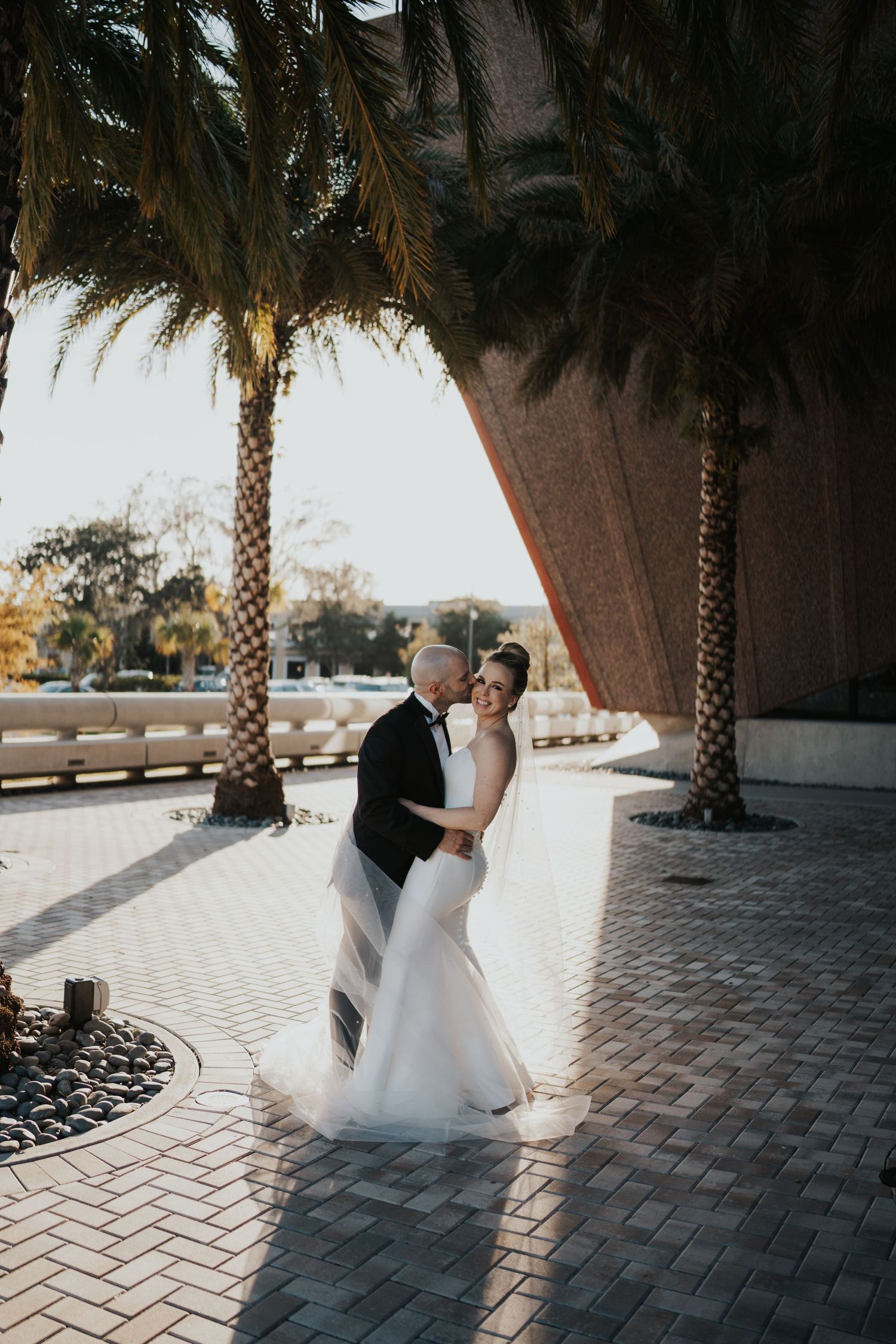 An elegant couple shares a romantic moment under palm trees and modern architecture.