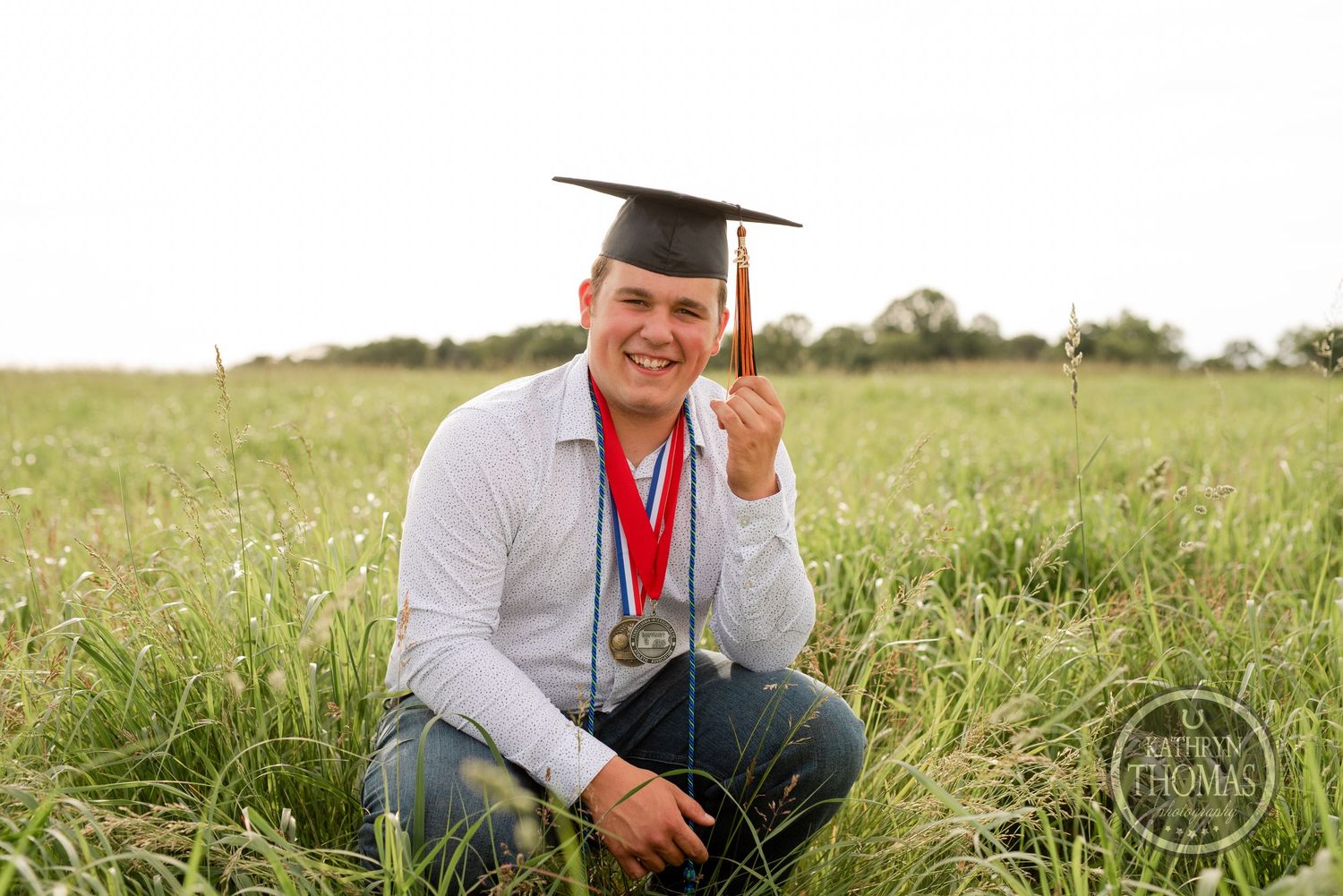 Graduate in cap and gown kneels in sunlit field during graduation photoshoot.