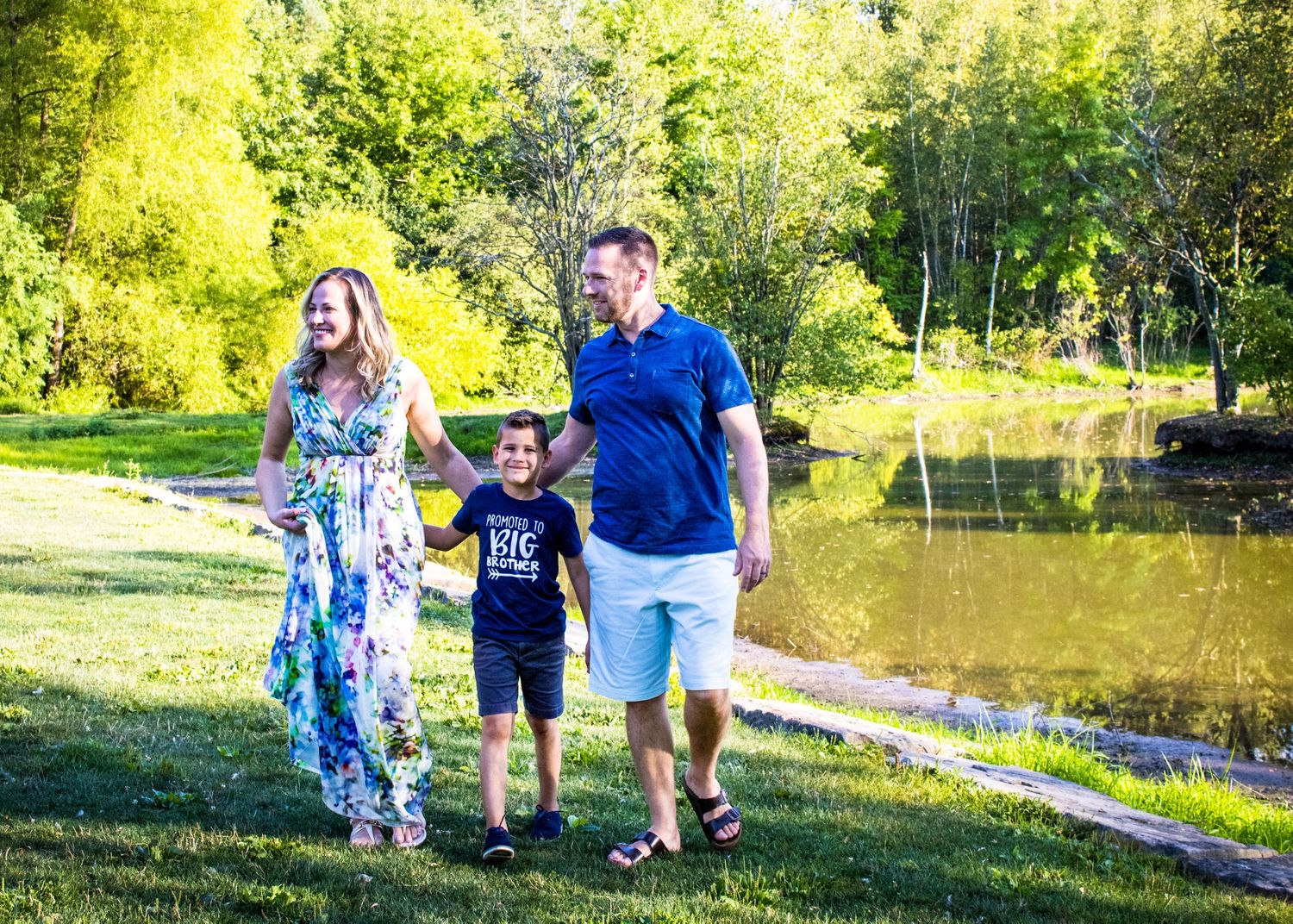 A mom, dad and a 7 year old boy walking arm in arm by a pond on a sunny, spring day.