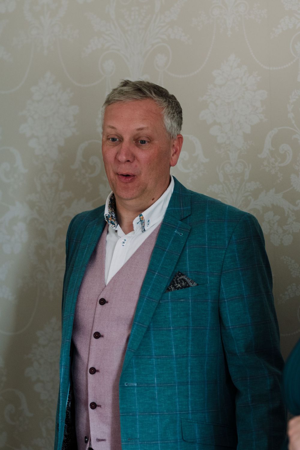 Person wearing a stylish teal blazer and pink vest against a wallpapered background.