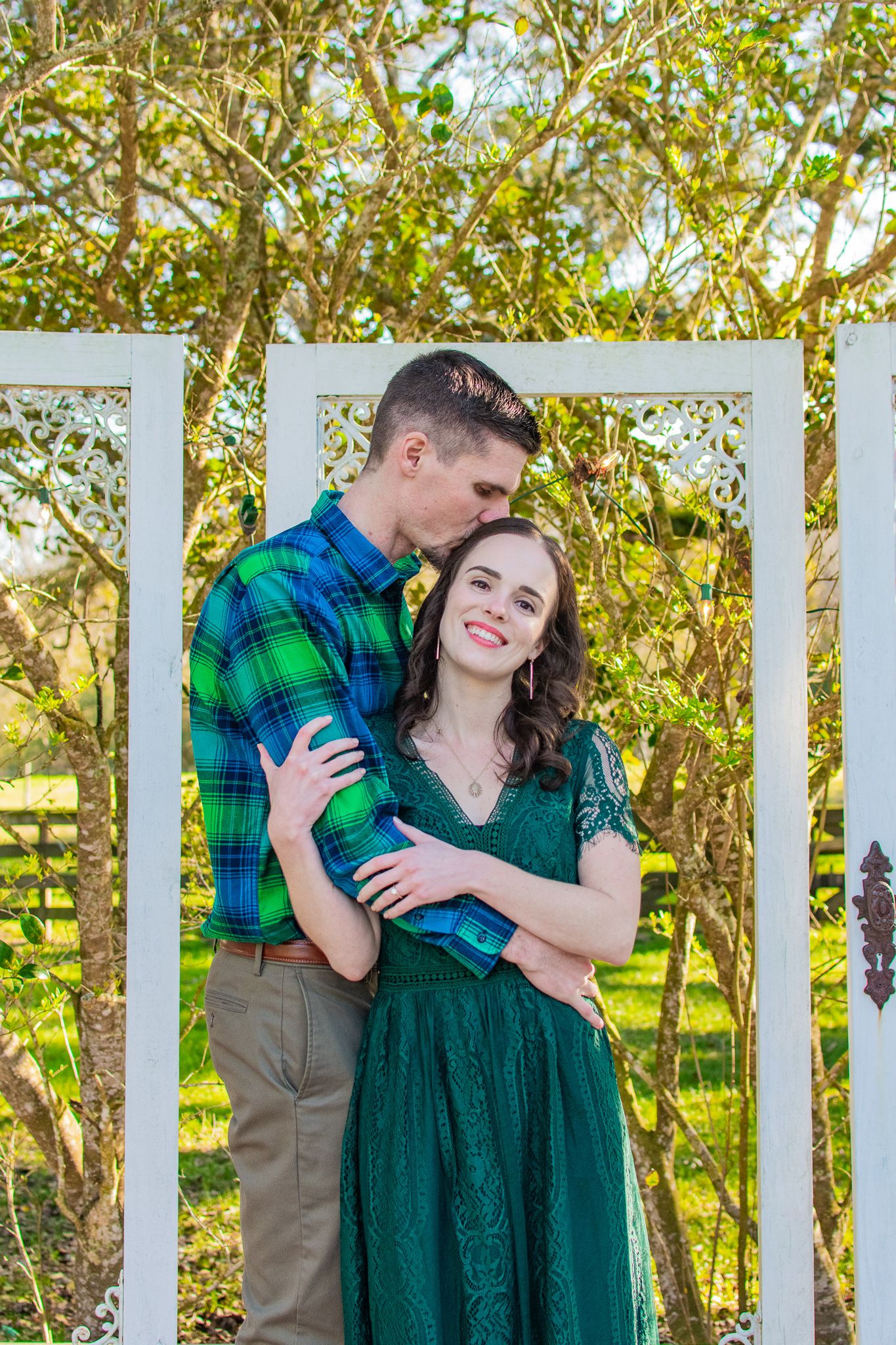 Couple shares romantic moment in front of white garden trellis surrounded by green foliage.