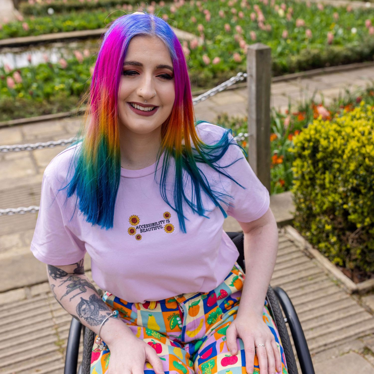 London rainbow haired disabled influencer posing sat smiling in their wheelchair wearing a rainbow outfit.