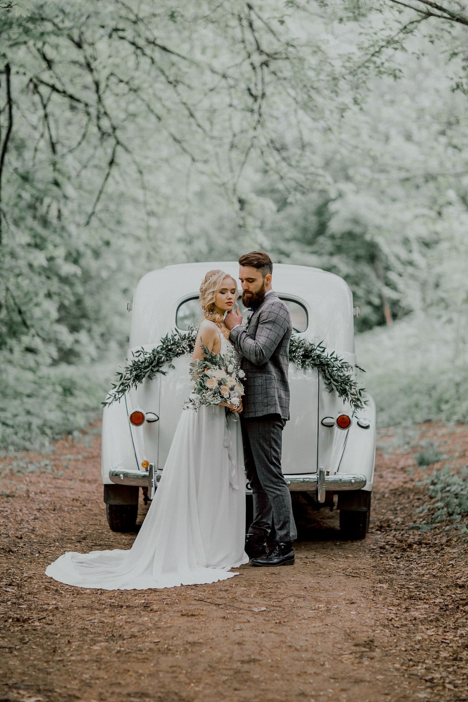 A couple poses romantically in front of a vintage white car decorated with greenery in a dreamy forest setting.
