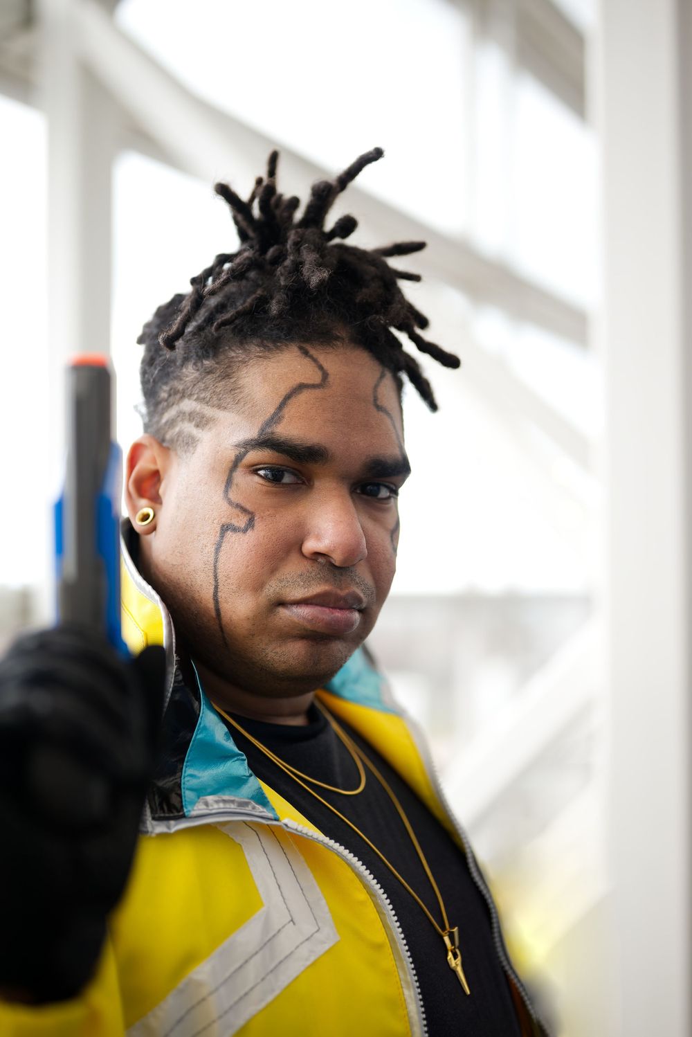 Man with dreadlocks and face tattoo in yellow jacket pointing a toy gun at camera in cosplay costume.