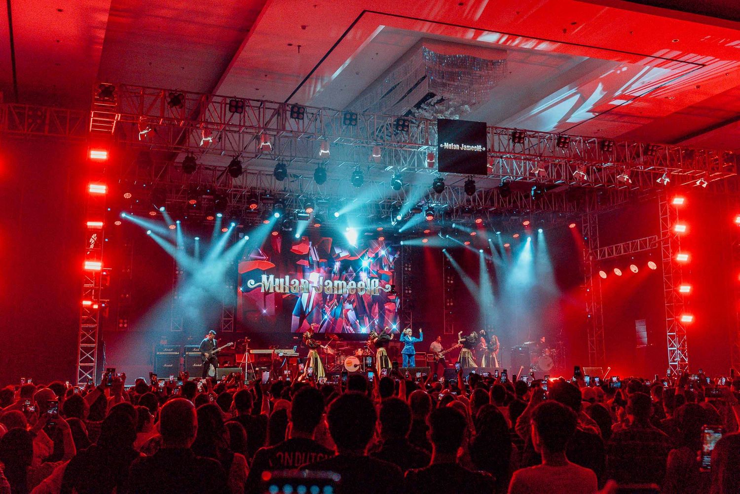 Concert venue filled with crowd silhouettes under dramatic red and blue stage lighting.