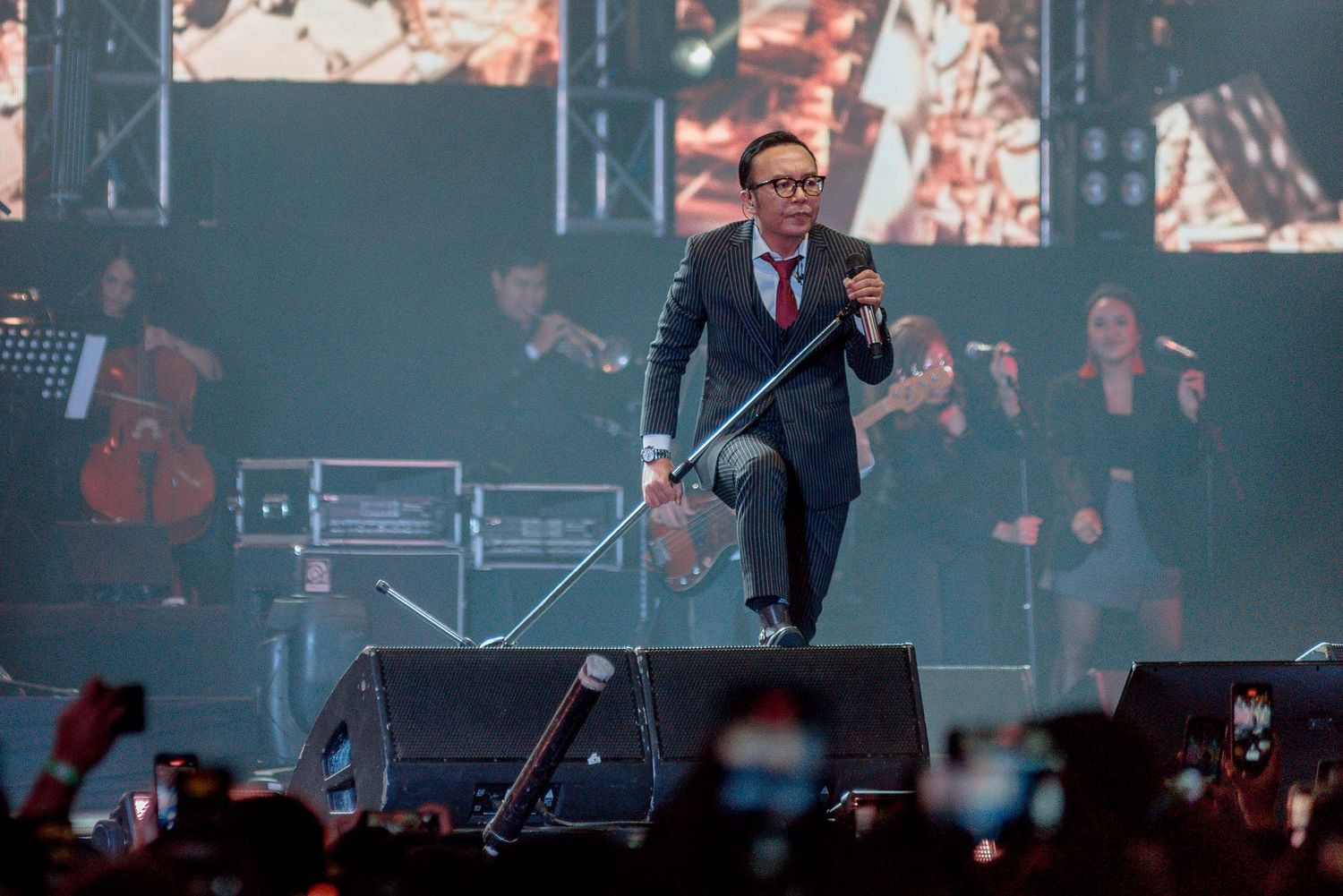 A performer in a suit leaps energetically on stage during a concert with musicians visible in the background.