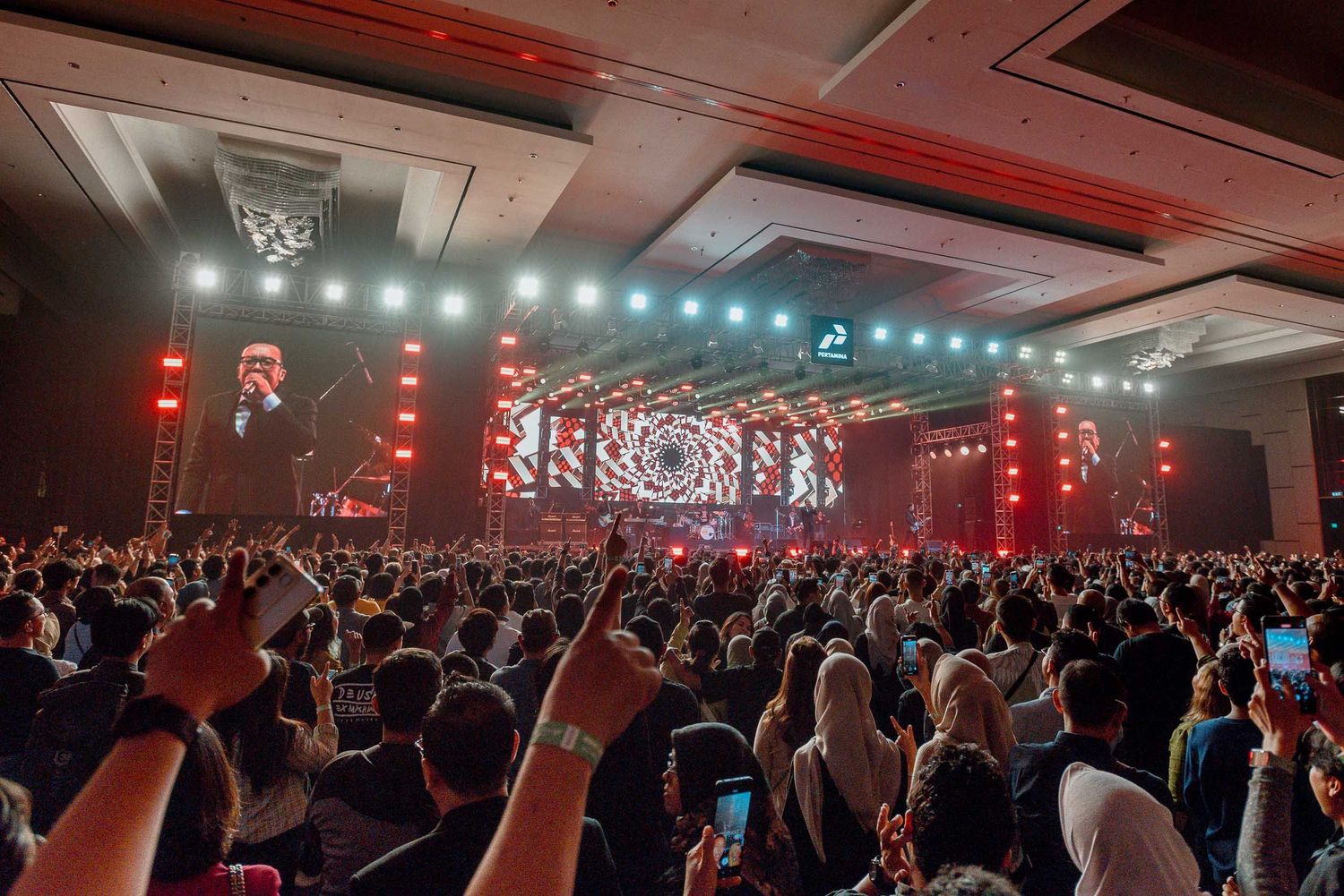 A packed concert venue with red lighting and crowd holding phones up during performance.