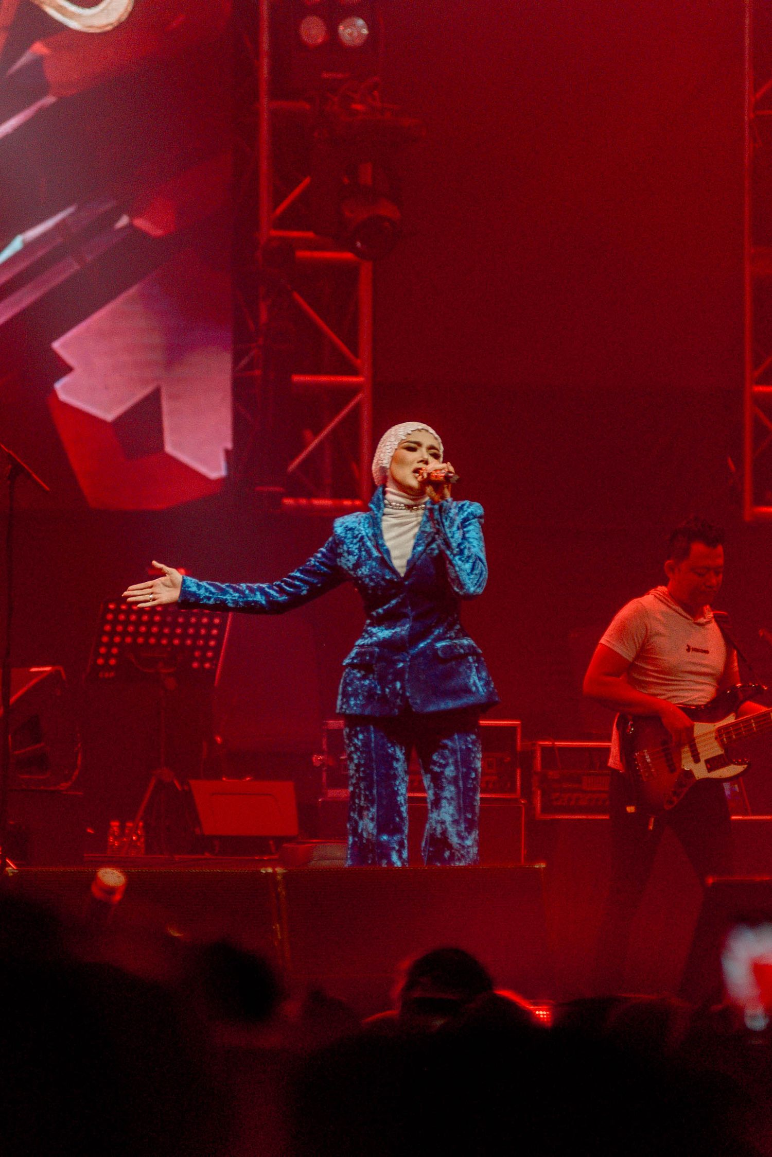 Performer in blue outfit singing on stage with red lighting and band members in the background.