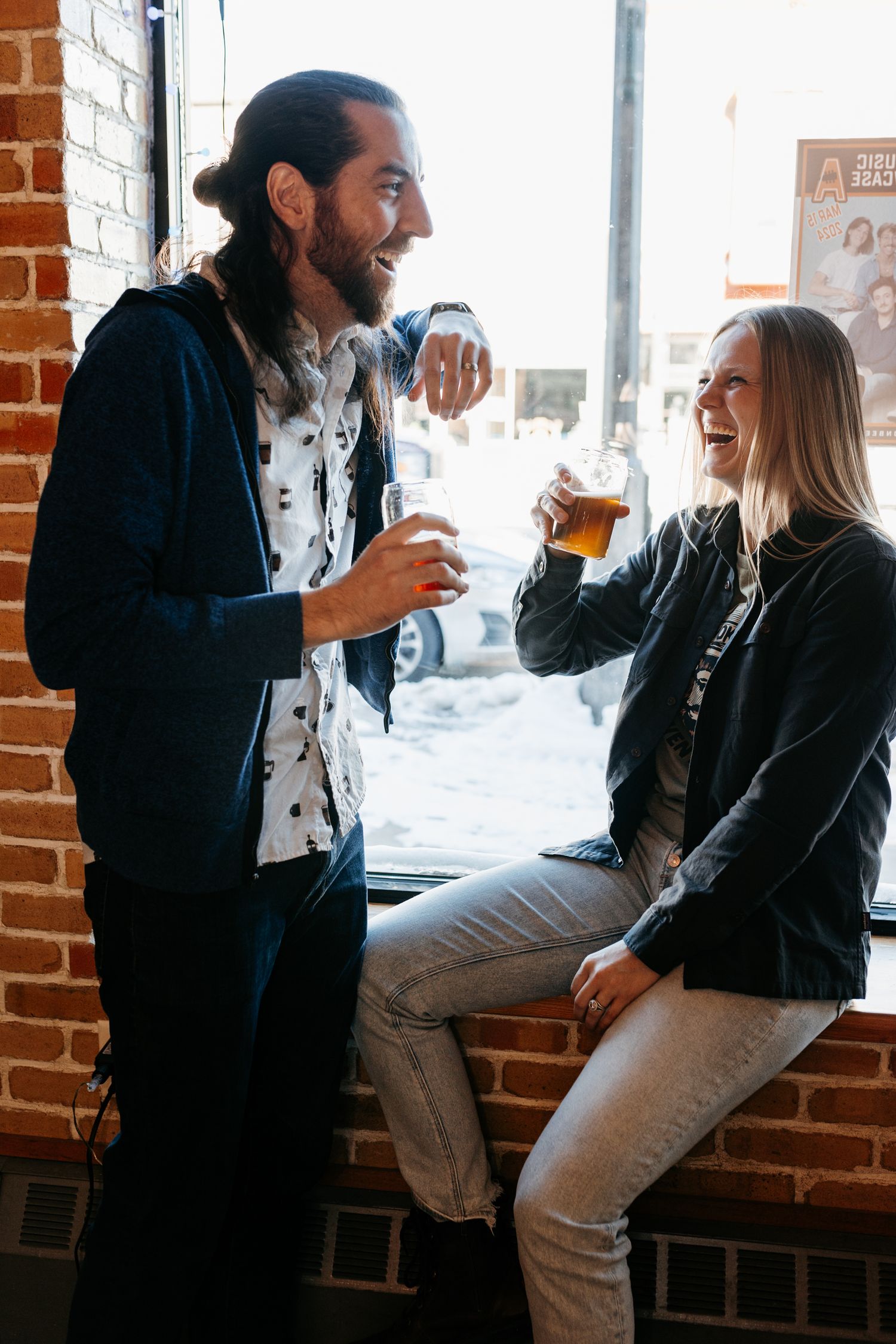 Two people share drinks and laughter by a brick wall in a brewery taproom.