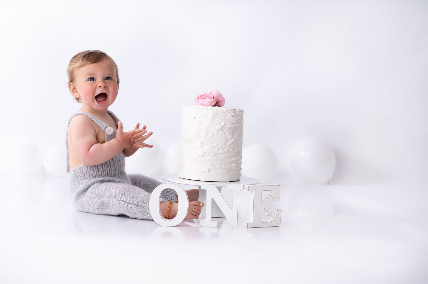 Baby sitting next to white cake and 'ONE' letters in studio setting for first birthday photoshoot.