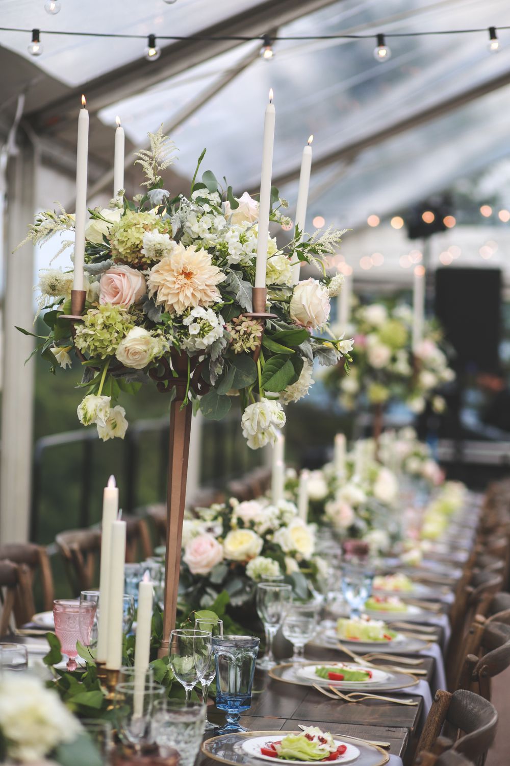An elegant wedding table setting with tall floral centerpieces, candles, and soft lighting under a clear tent.
