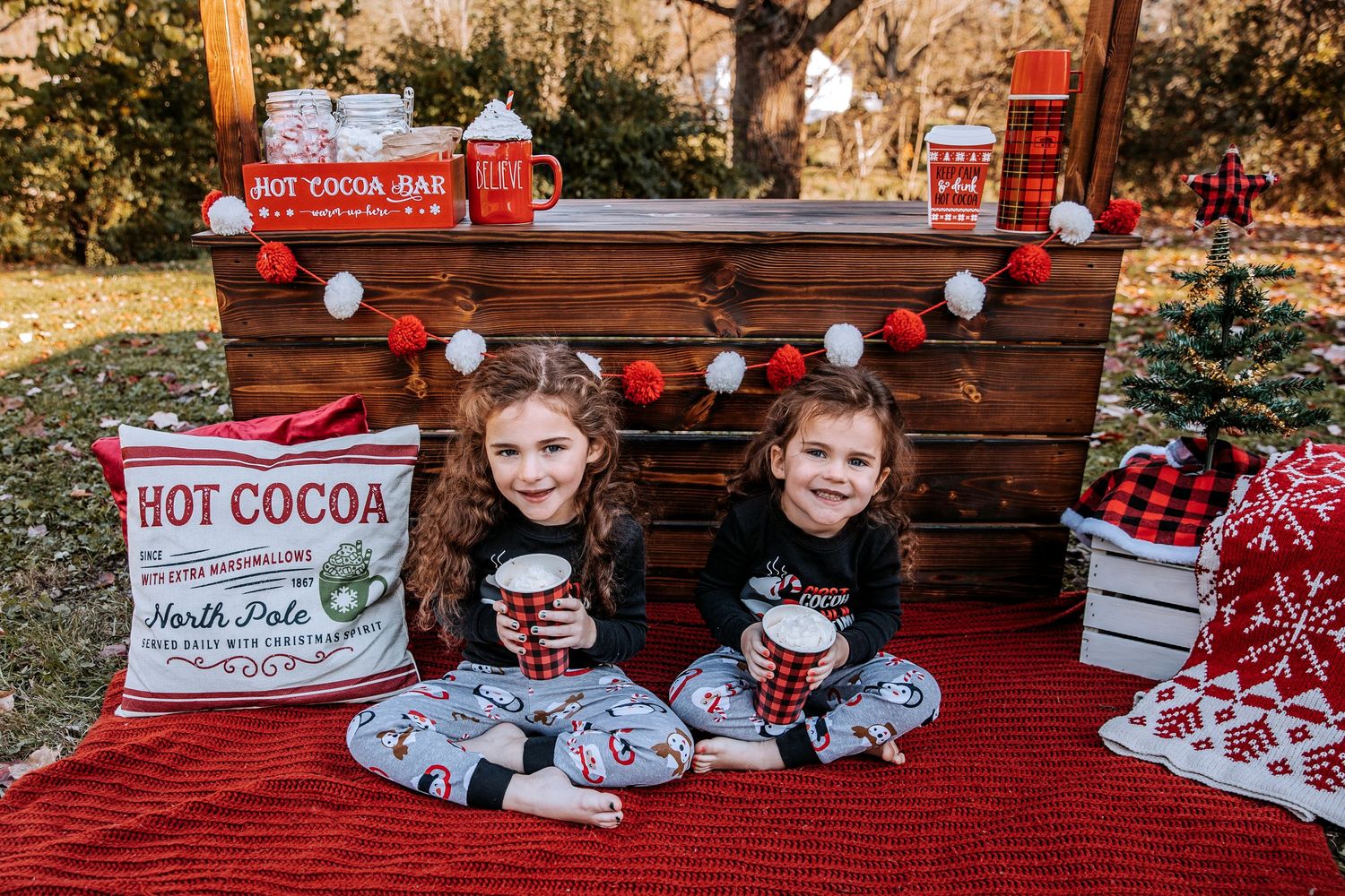 Two children sit on a red blanket with hot cocoa decorations and wooden backdrop for holiday photos.